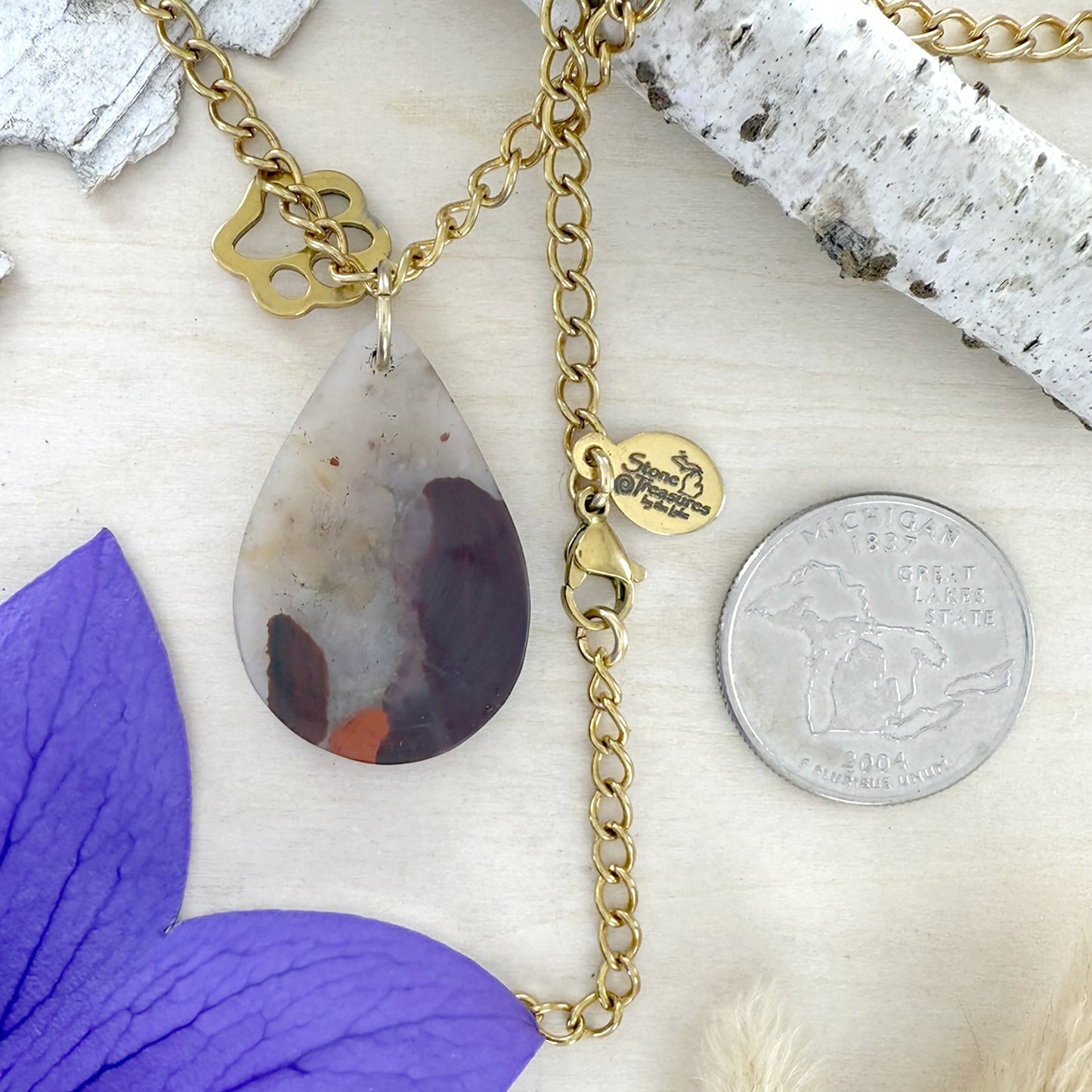 Gold necklace with a teardrop-shaped pendant on a white surface with a purple flower and silver coin for scale.