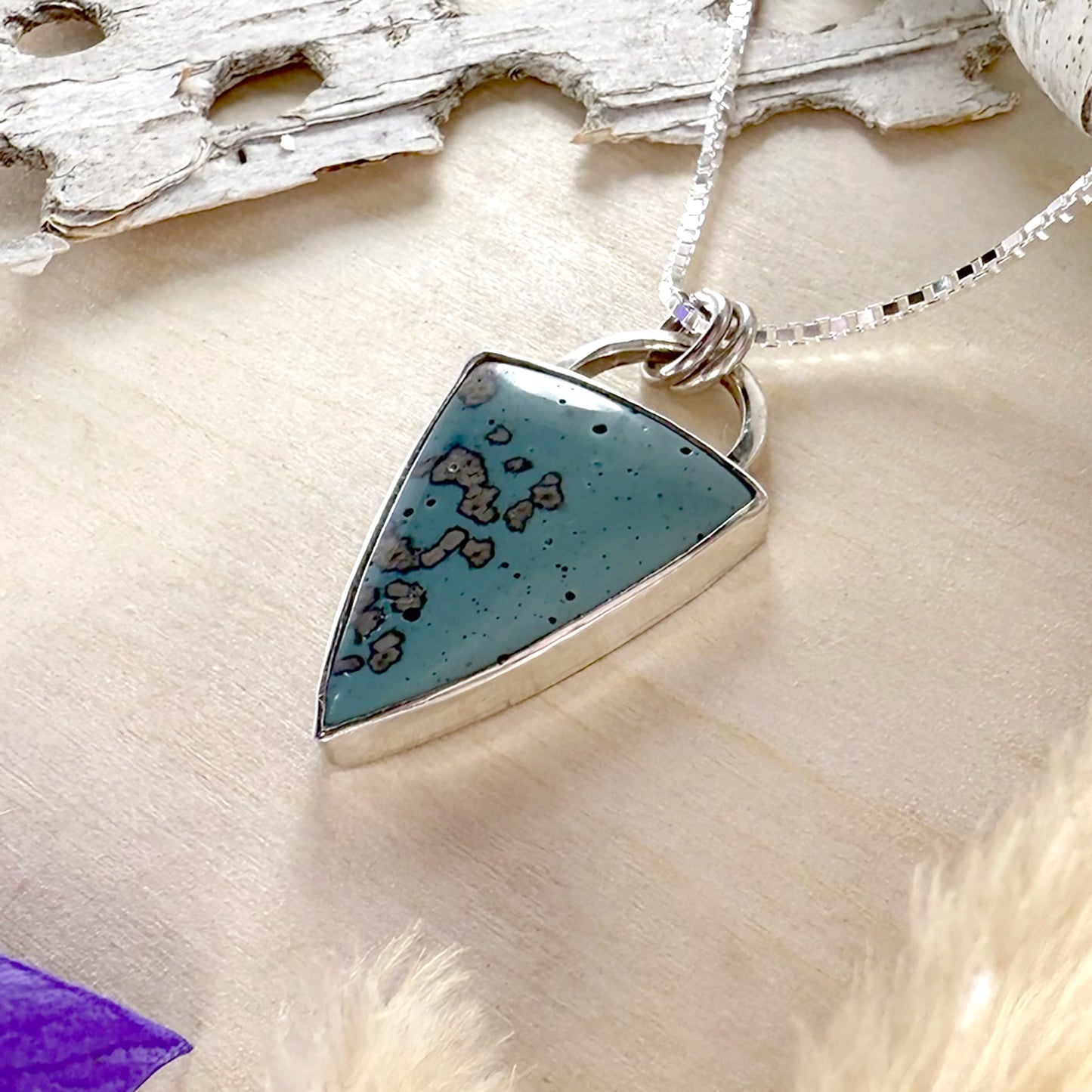 Triangular pendant with speckled design on a chain, set against a natural wood and shell background.