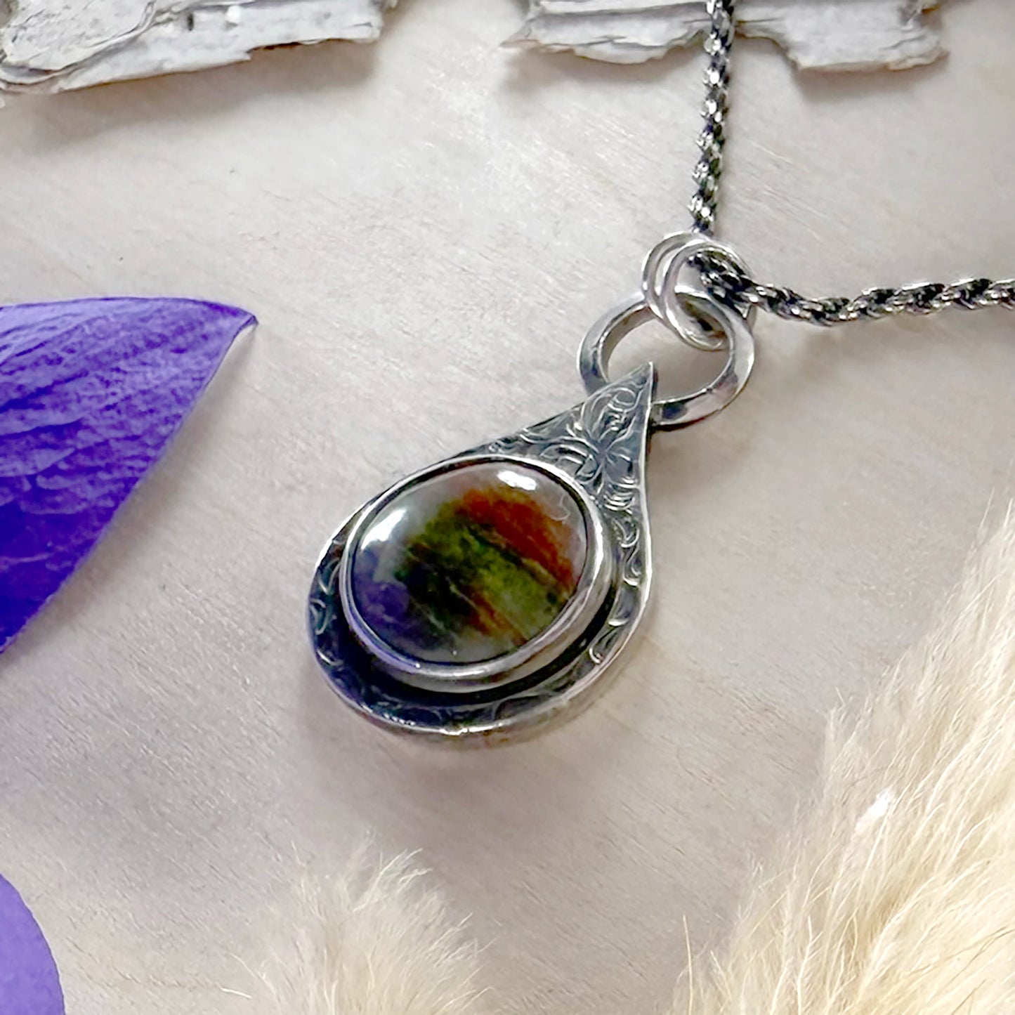 Silver pendant with a colorful stone on a wood surface with purple and white elements.