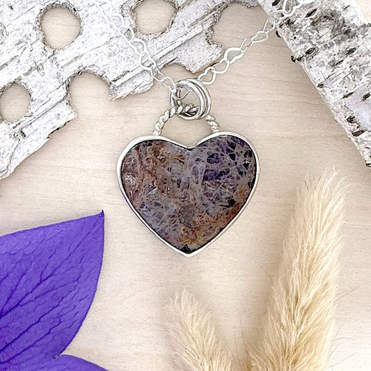 Heart-shaped pendant with a natural stone design on a chain, placed on a textured surface with a purple leaf and dried grasses.
