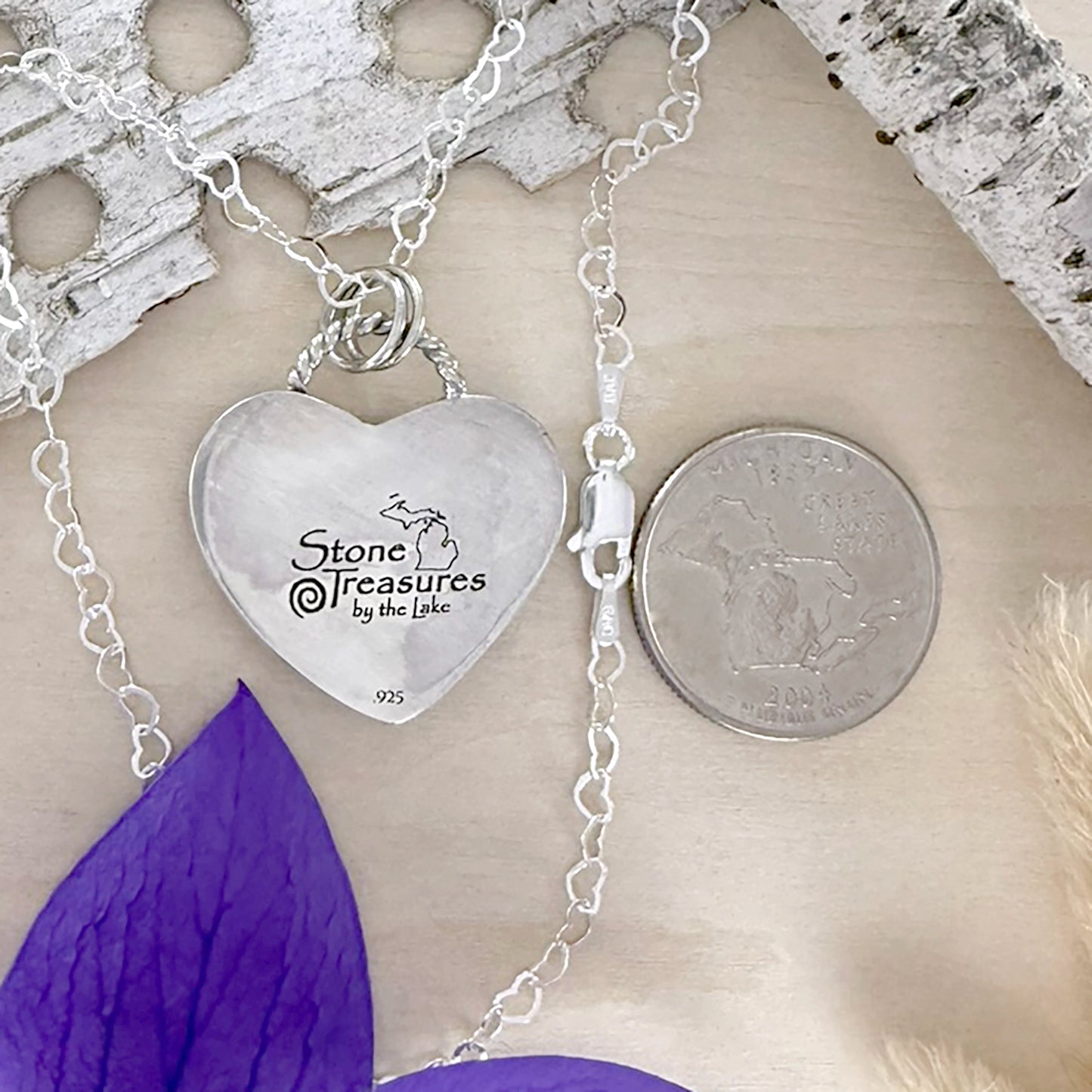 Heart-shaped silver pendant with 'Stone Treasures by the Lake' engraving on a chain, next to a quarter for scale.