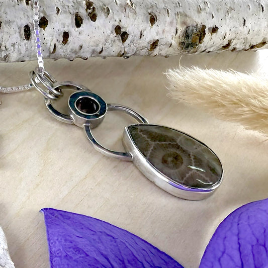 Silver pendant with a teardrop-shaped stone on a textured surface with purple flowers.