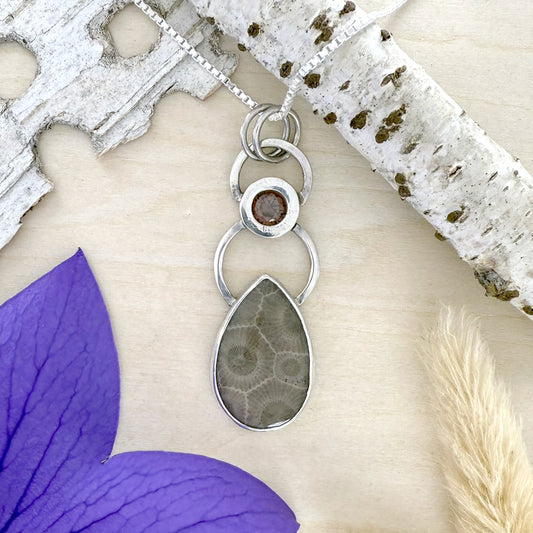 Silver pendant necklace with a teardrop-shaped stone on a white surface with purple flower and dried plant.
