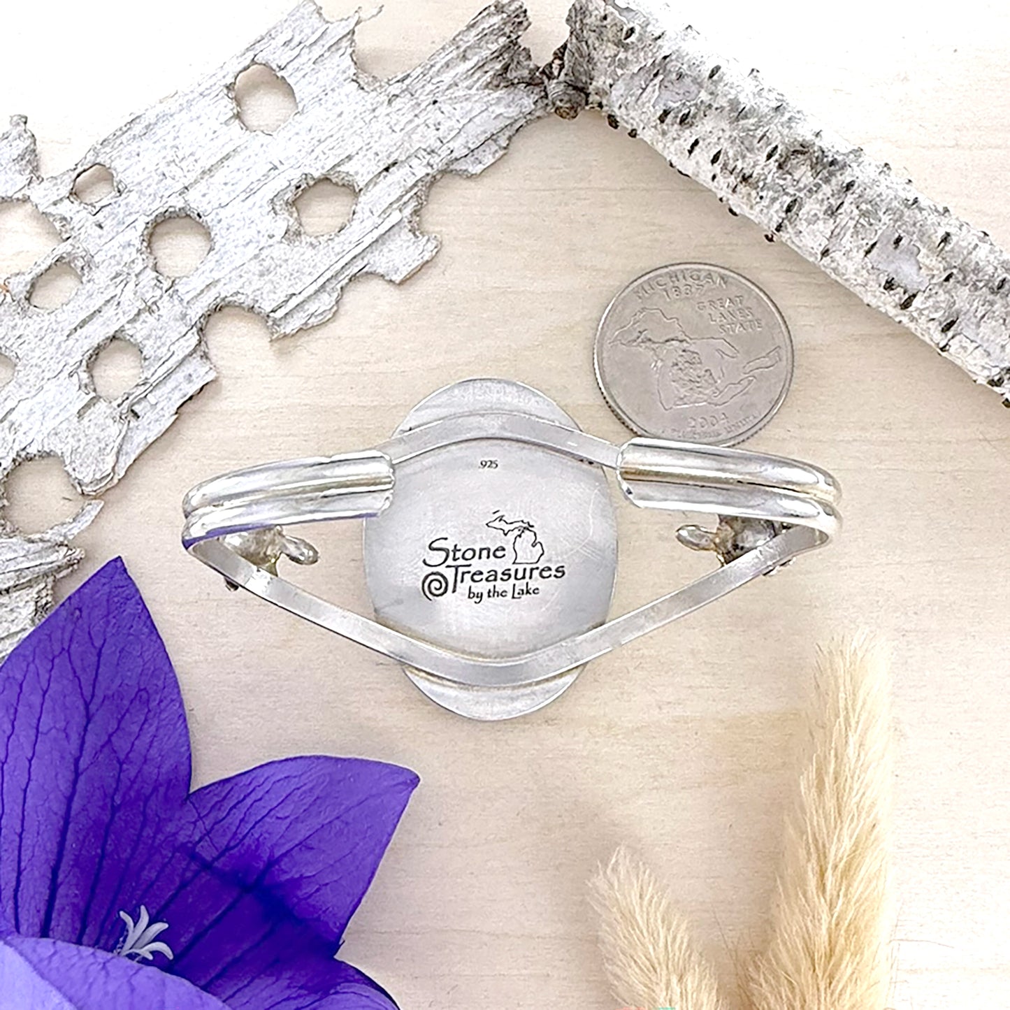 Silver bracelet with 'Stone Treasures by the Lake' engraving with a purple flower and silver leaf accents.