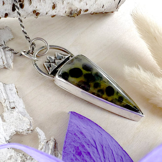 Silver pendant with a green and black speckled stone on a wood surface.