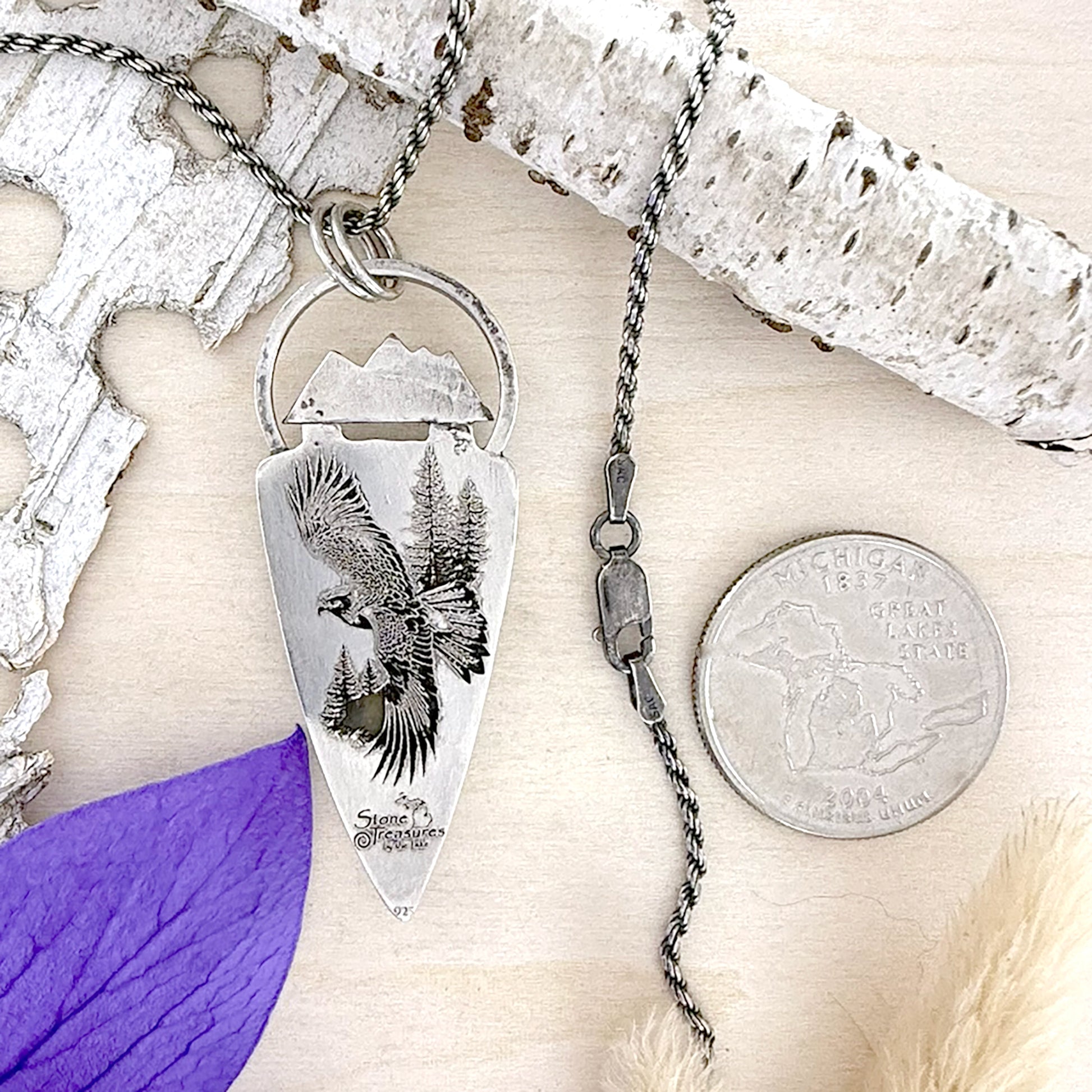 Silver pendant with engraved design on a wood surface, next to a coin for scale.