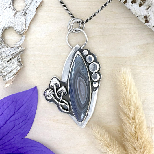 Silver pendant with a natural stone design on a textured surface with purple flower and dried plant elements.