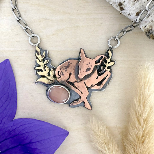 Necklace with a deer pendant on a textured surface with purple flower and beige plant.