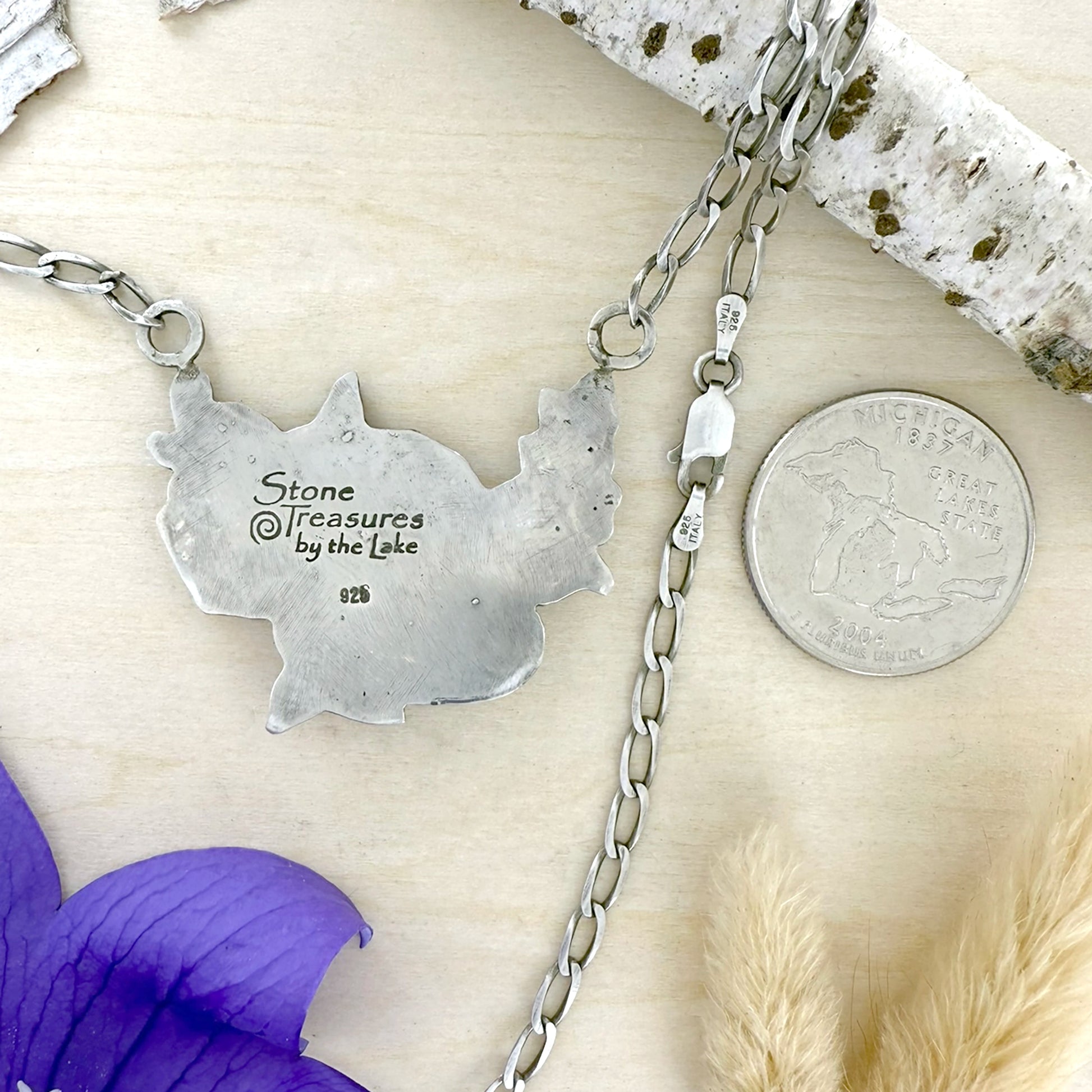 Silver necklace with a pendant and chain on a textured surface with a coin and purple flower.
