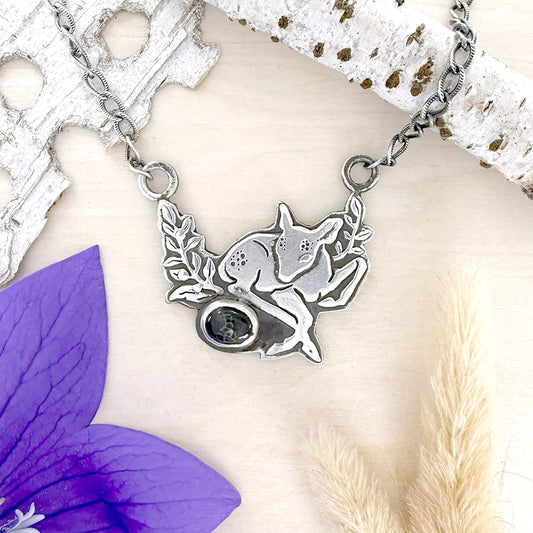Silver fawn necklace with a gemstone on a white surface with purple flower and beige grass.